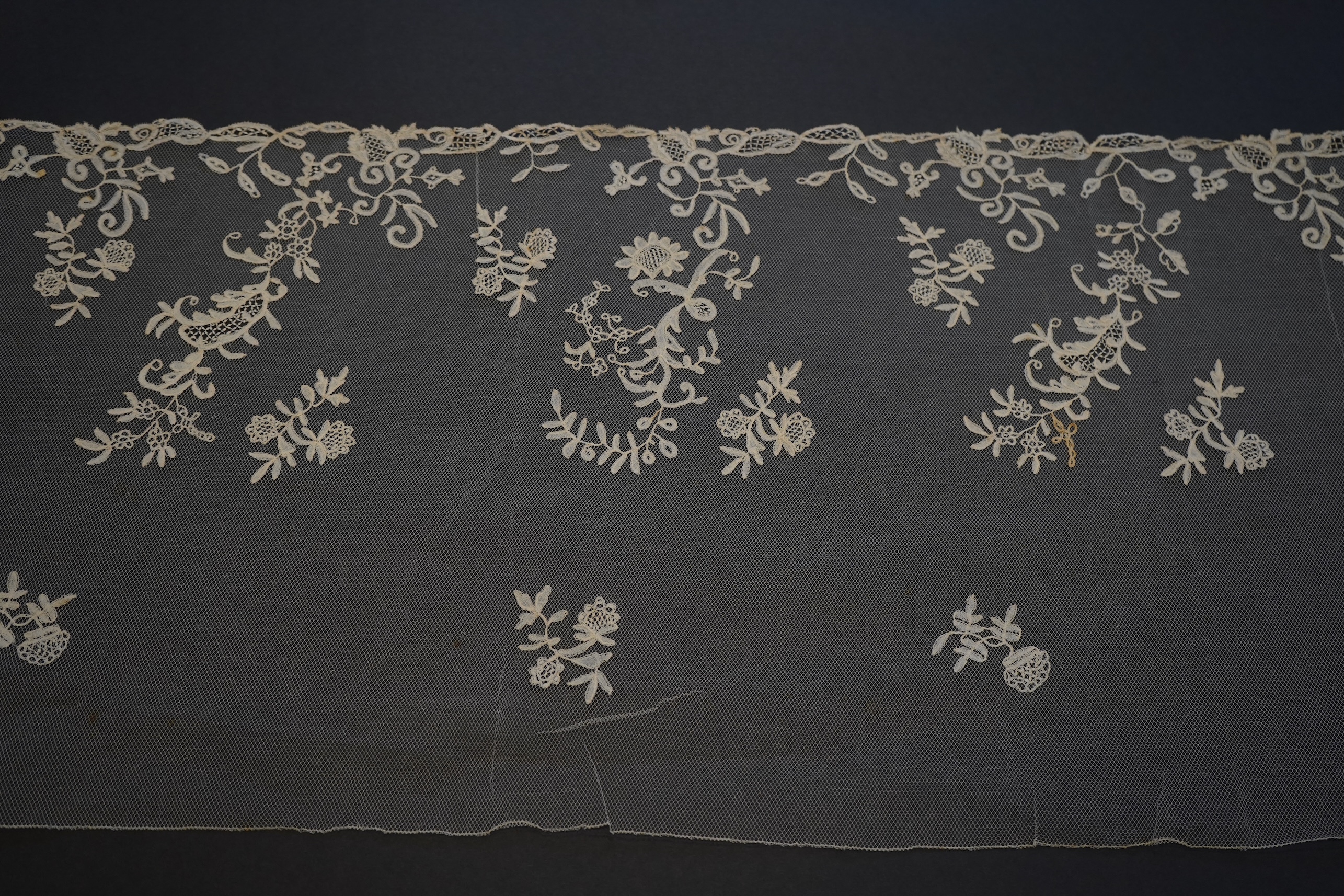 A 19th century five yard flounce of Brussels bobbin appliqué lace applied onto fine machine net, worked in a trailing floral design with an ornate border and sprig motifs, 457.5 cm long x 36cm deep. Condition - possibly